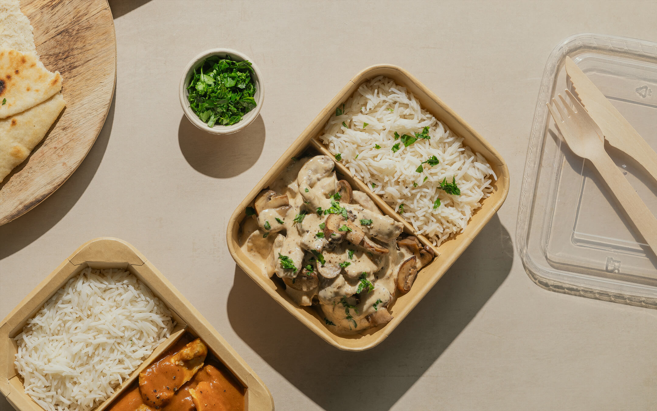 Display of food delivery kraft packaging, wood cutlery and plastic lid packed with rice, chicken and creamy mushroom sauce.